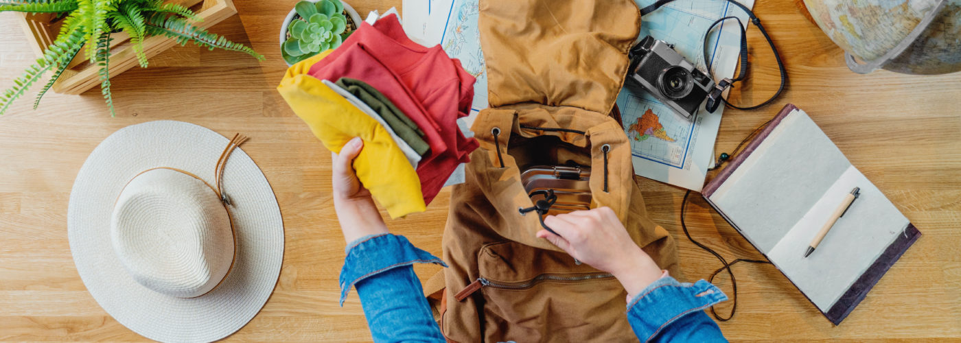 Aerial view of person packing backpack surrounded by maps, a camera, a globe, and a hat