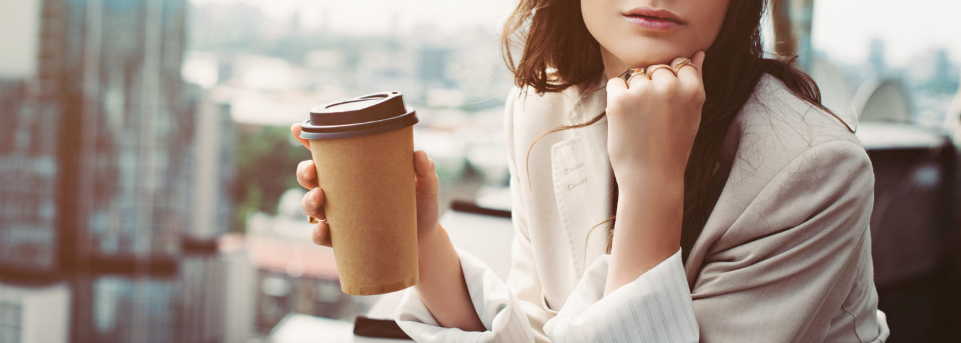 Woman holding a to-go coffee cup in front of a large glass window with view of the city