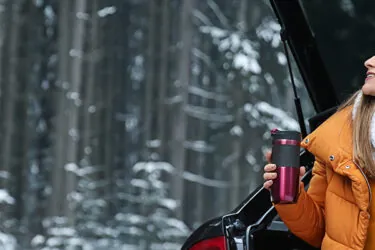 Woman sitting in open trunk of car, drinking coffee in the snow